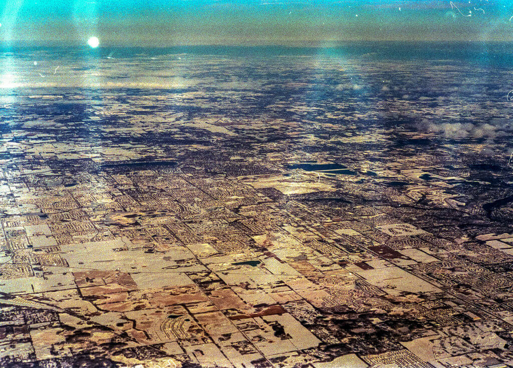 From above, the expanse of land unfolds like a meticulously woven quilt, each patch a testimony to nature's quiet symmetry. Snow blankets every inch, its pristine whiteness interrupted only by the faint demarcations of patchwork fields below. The horizon draws a delicate line beneath an overcast sky, clouds assembling in unhurried clusters that hint at stories untold. Sunlight filters through intermittently, casting sporadic glares that intrude upon the camera’s lens—an unwanted but persistent guest in the upper left corner of this captured moment. Here and there, where light penetrates the cloud cover with just enough insistence, reflections shimmer and dance briefly on this wintry canvas before giving way once more to softer shades and muted tones.