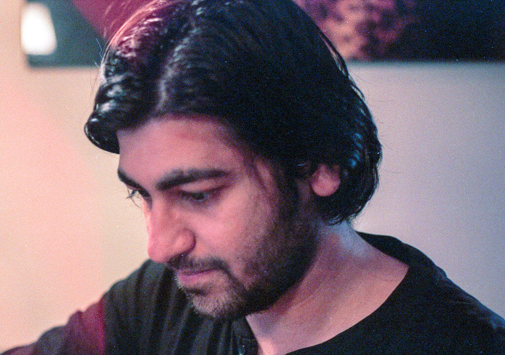In a room suffused with the tender embrace of warm light, a man stood, his medium-length dark hair falling gently over his forehead. His beard, meticulously grown and hinting at days spent in quiet introspection, framed his face. Clad in a dark shirt that seemed to absorb the softness of the surroundings, he gazed downward, lost in thought. The world beyond him blurred into subtle shades of warmth, an indistinct watercolor that softened the edges of reality. The light caught his hair just so, casting fleeting glimmers that seemed to echo his reflective mood—a portrait not just of a man but of contemplation itself.