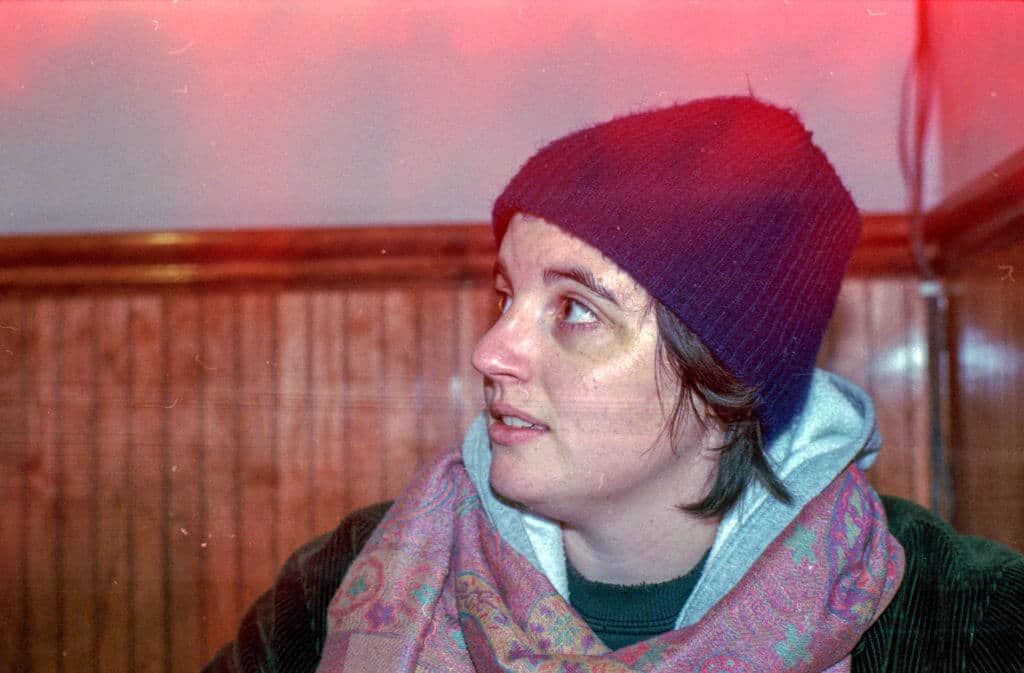 The individual, cocooned in a cozy assemblage of attire—a navy blue beanie snug atop their head, a pink scarf gracefully swathed around their neck, and a gray hoodie offering an extra layer of comfort—gazes slightly upward to the left. They are ensconced within the intimate confines of an indoor space, where the warmth of wooden paneling serves as the backdrop. Overhead, an unexpected red light leak spills across the photograph like spilled ink on parchment, imbuing it with a sense of nostalgia and quiet reverie.