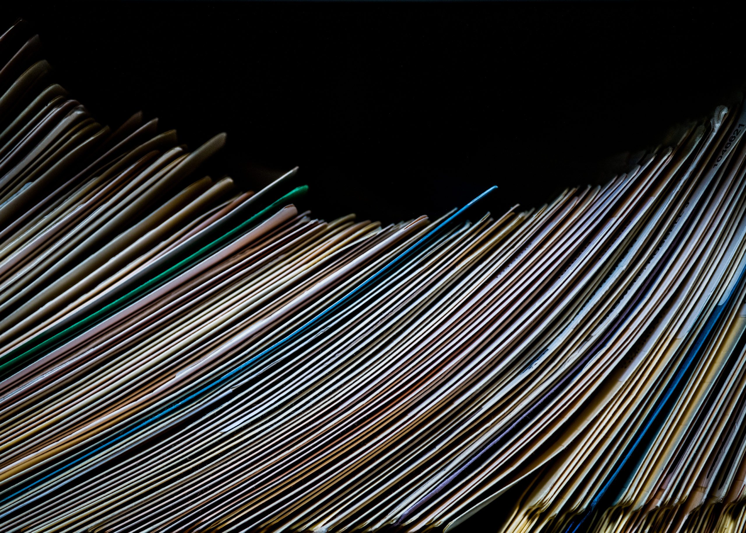 The scene captures an intimate tableau of countless documents, meticulously arranged in a gentle, undulating curve. Each paper, with its own distinct hue of cream, tan, and light brown, tells a silent story of age and weathering. Edges curl ever so slightly, whispering secrets of times past. The dark background frames this assembly with stark contrast, giving the papers an almost reverential prominence as if they exist in their own delicate universe.