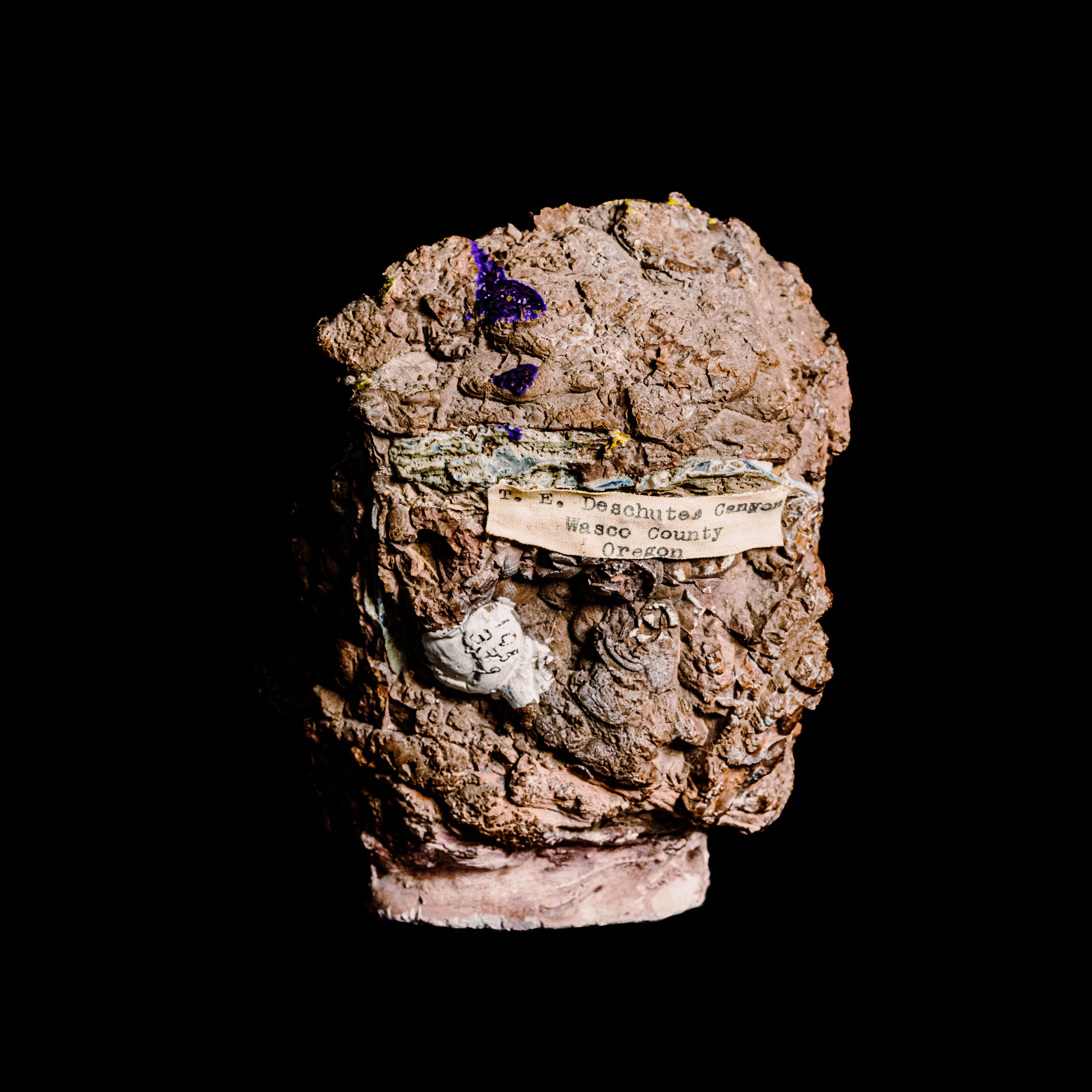 Deen-Sanwal_OMSI-85 Set against an obsidian background, the mineral specimen's rugged texture immediately catches the eye. Its earthy, brown hues evoke a sense of ancient terrain, whispering stories of its long journey through time. A slightly crumpled and yellowed label clings to its side, hinting at origins rooted deep within Deschutes County, Oregon. The inscription is partially obscured by time's embrace but still offers enough detail to connect the past with the present. The display creates a silent recollection of place and history, both enduring and fragile in their coexistence.
