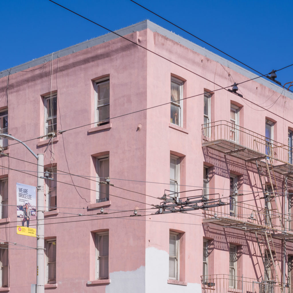 The building, washed in a shade of pink reminiscent of rose petals, stood tall and resolute against the expanse of the sky, its four stories punctuated by windows that seemed to peer curiously at the world below. A fire escape clung to one side like an afterthought, a pragmatic addition rendered almost poetic by its stark contrast against the serene blue above. 

Intricate webs of power lines crisscrossed in front, their presence both intrusive and comforting—veins carrying lifeblood through the city. On a nearby utility pole at the left corner, a poster flapped gently in the breeze. It bore a simple message: "We Are Service Net." The words were imbued with an earnest promise that hinted at connection and reliance in this bustling urban tableau.