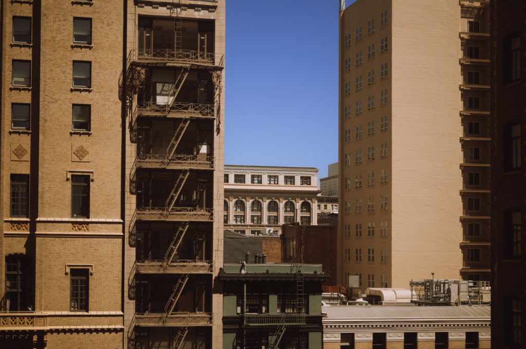 The cityscape unfolds like a meticulously composed painting, dominated by a cluster of tall, beige buildings. Each structure stands resolute, adorned with steady fire escapes that cascade downward, their iron frames casting intricate shadows onto the masonry. The large windows punctuate the facades with a regular rhythm, reflecting glimmers of lives unseen within.

In the distance, a white and grey building rises dignified and distinct against the crisp azure expanse of sky. It stands framed by its foreground counterparts, an architectural island serenely anchored amidst the urban tide. The clear brightness above lends an air of tranquil clarity to the scene; everything rendered in sharp relief yet partaking in an implicit harmony—an interwoven existence sustained under an ever-watchful blue canopy.