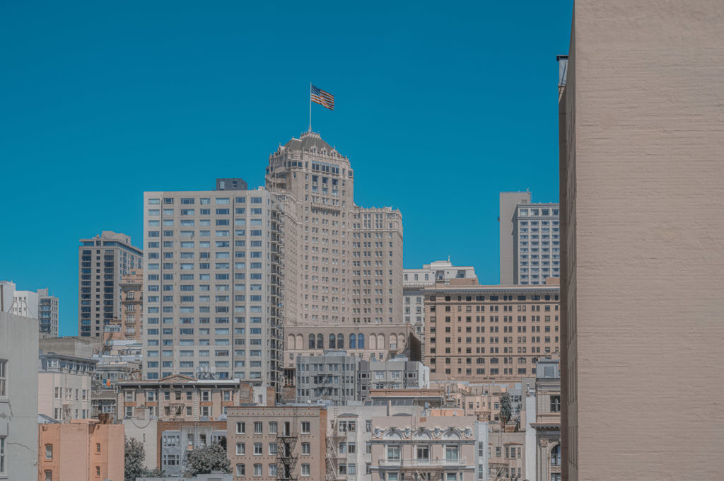 On this crisp morning, under the embrace of an unblemished blue sky, the city reveals itself with a serene majesty. A dignified building, crowned with an American flag that flutters gently in the breeze, stands as a sentinel amidst a multitude of structures. These edifices rise and meld into one another, forming a complex tapestry of urban life. From my vantage point, I see how beige and light-colored facades dominate both foreground and horizon, each building an architectural story etched against the azure expanse above. Here amongst these towering forms is where lives converge and diverge in silent symphony, shaped by the rhythm of their interplay within this sprawling mosaic called home.