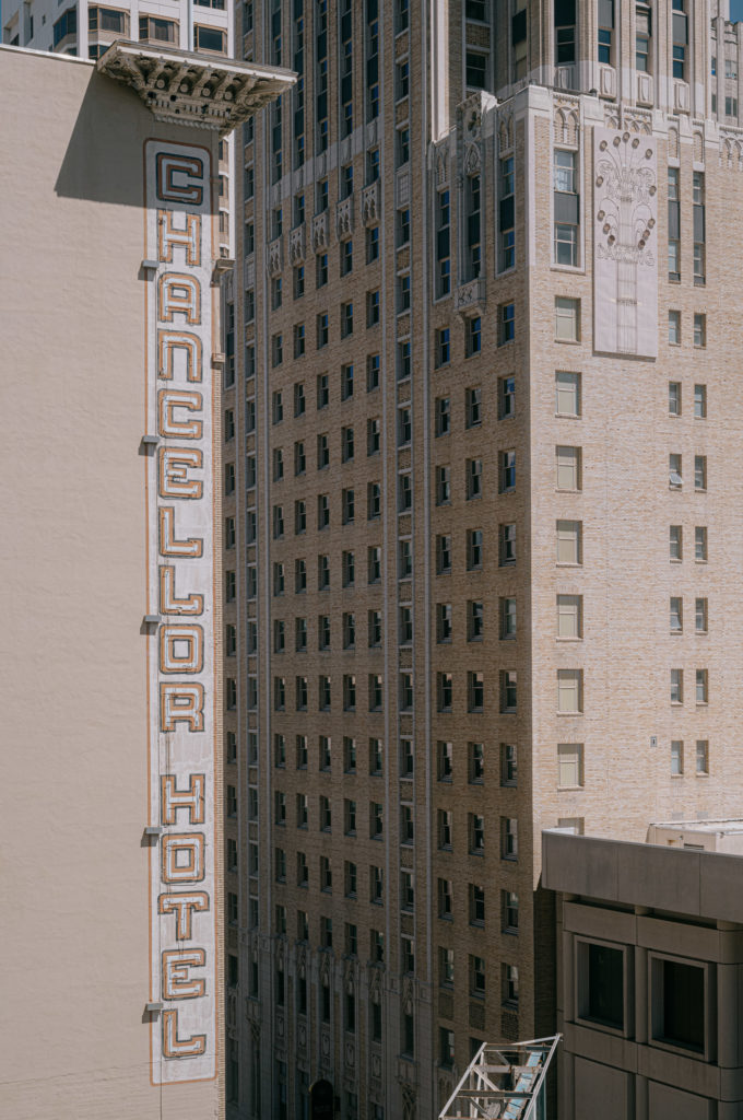 The Chancellor Hotel, its signage a testament to the building’s prominence, stood unwavering and tall in the cityscape. The bold letters of its name reached skyward, embodying both history and grandeur. Behind it, a mosaic of light-brown high-rises rose to different heights and angles, each adorned with windows that seemed like eyes into countless stories. Ornate moldings traced their edges, adding an old-world charm to their facades. A beige building in the ensemble drew attention with its intricate details and a partially visible banner that whispered tales of bygone eras amidst the urban hustle.
