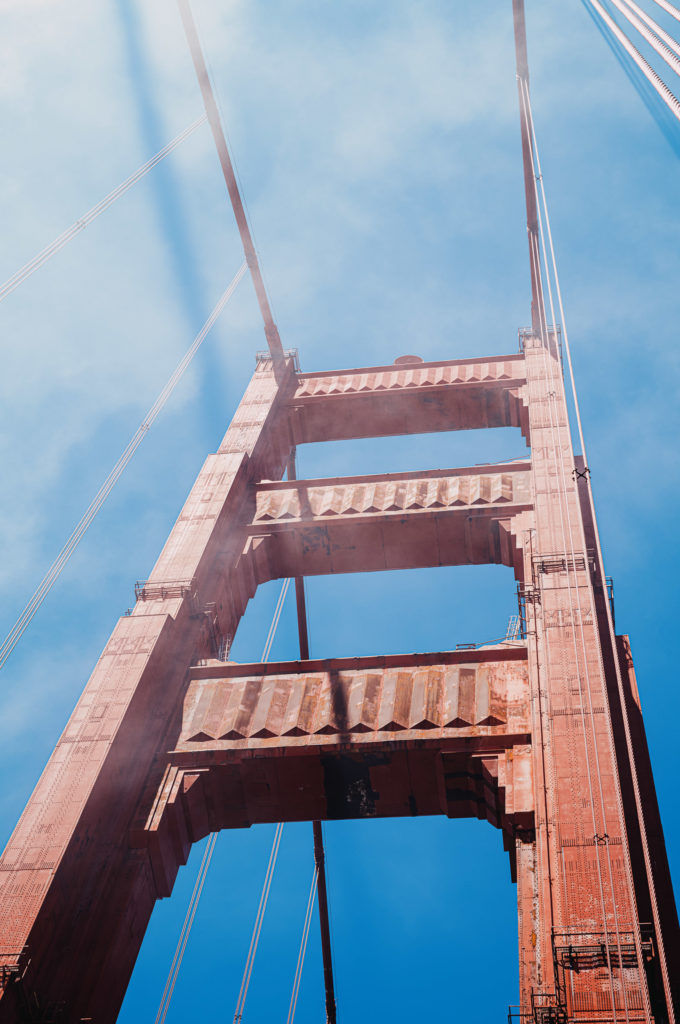 From a vantage point below, the iconic crimson tower of the Golden Gate Bridge in San Francisco soars upwards, an omnipresent sentinel against the expanse of a cloudless blue sky. There is a haze—subtle yet pervasive—that imbues the scene with a fleeting sense of impermanence. The bridge's sinewy cables fan out above like delicate threads, weaving through air and mist alike. This perspective, grounded yet aspirational, underscores the monumentality of the structure—a conduit between worlds mundane and sublime. The bridge stands resilient against time and tide, its steadfastness mirrored in every rivet and beam that holds it together.