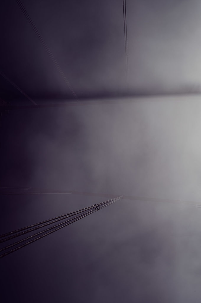 A brooding, melancholic scene emerges as the sinewy cables of the bridge extend into an ethereal fog. The thick mist, a relentless shroud, obscures the familiar contours of the structure, rendering it almost spectral. As these steel tendrils vanish into the grayscale abyss above, they invoke a sense of haunting beauty—an atmospheric dance between man-made certainties and nature's elusive ambiguities. This tableau speaks in whispers to those who pause to witness its somber grace.