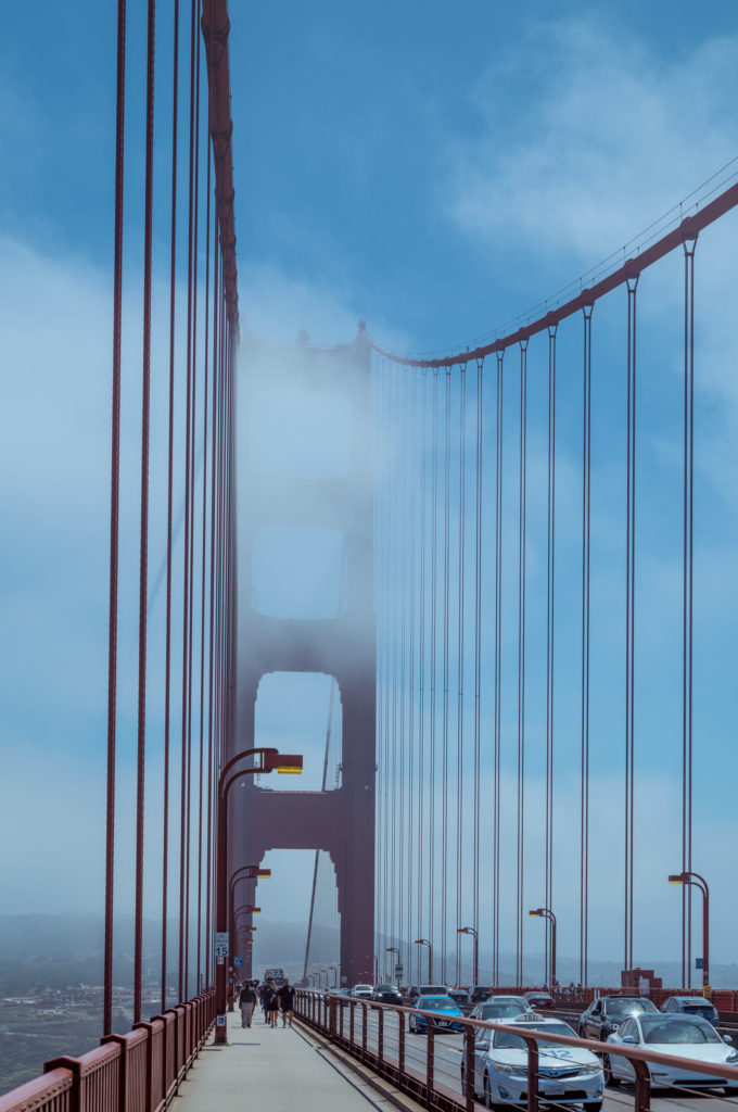 Through the mist that clung to the morning, the Golden Gate Bridge emerged in fragments, its formidable red-orange towers standing resolute against a backdrop of relentless greyness. The suspension cables stretched like taut threads of fate, vanishing into the fog only to reappear farther along their destined path. On one side, a procession of pedestrians meandered along the walkway, their silhouettes mingling with an air of quiet anticipation—a silent communion with this iconic passageway. On the other side, vehicles coursed steadily on the roadway, their purpose clear yet subdued by the enveloping vapor that dulled all perceptions but heightened every sensation. It was an everyday pilgrimage for some and a moment of awe for others—a microcosm suspended between solidity and ethereal drift.