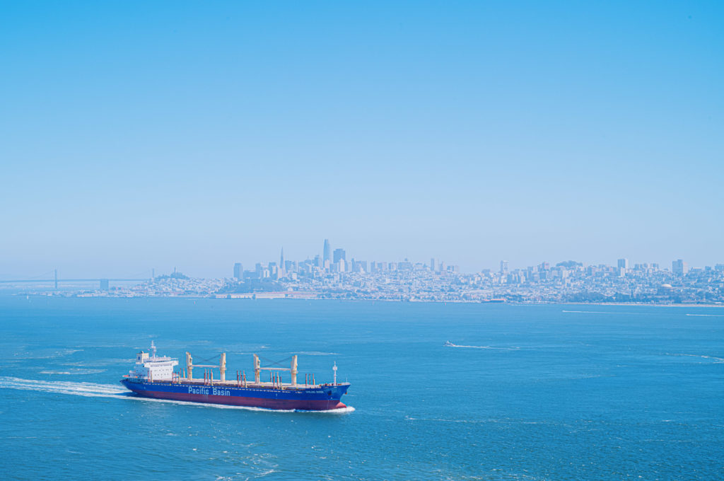 The cargo ship bore the name "Pacific Basin," its hull a muted, steadfast presence against the expanse of an endless, cerulean sea. As it cut through the water with determined grace, the silhouette of a distant city appeared—its skyline softened by a tender haze. Towering high-rises stood like sentinels beside an intricate bridge that arched over unseen currents. Above, a sky stretched in flawless serenity, casting its unwavering gaze on the convergence of industry and nature below. In this tranquil moment, the vastness of both the ocean and sky seemed to hold secrets beyond time and distance.