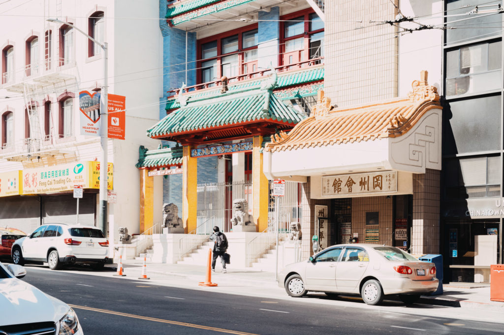 Amid the rhythm of city life, a building adorned with traditional Chinese architectural motifs stands as a testament to heritage and history. Its ornate rooflines, resplendent in hues of red and green, curve gracefully under the weight of time. Two stone lion statues, resolute and guardian-like, flank the entrance with a quiet dignity. Alongside the street, everyday cars rest in casual disarray, their mundanity contrasting sharply with the structure's elegance. A solitary person walks near the entrance, seemingly unaware of their role in this tableau where past whispers gently entwine with present murmurs.