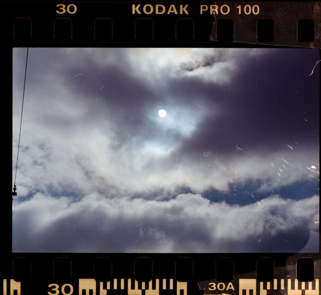 The photograph reveals a sky draped in clouds, the sun half-hidden, its presence more implied than seen, as if playing a coy game with the observer. Captured on Kodak PRO 100 film, the image bears an aura of nostalgia. The black borders of the film frame it with precision and numbered frames punctuate its edges like quiet sentinels. In the upper left corner, a thin cable snakes into view—a tangible thread in an otherwise ephemeral scene—reminding us of connections that tether unseen worlds together.