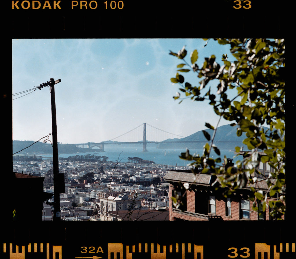 From a vantage point nestled within the neighborhood, the Golden Gate Bridge stretches elegantly across the bay, its iconic red hue captaining attention against an azure backdrop. The view, partially obscured by foliage on the right, frames this architectural marvel with a delicate intimacy. Below, San Francisco unfurls in a tapestry of rooftops and buildings, each structure telling its own story under the pristine sky. A filmstrip border edges this moment; "Kodak Pro 100," it reads—a silent testament to a captured instant in time.