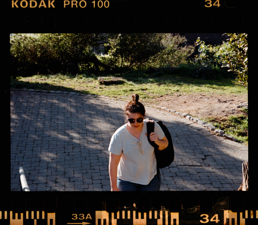 A figure ascended the cobblestone steps, their bun held meticulously, a small testament to their own morning ritual. The sun generously bathed the path, its warmth palpable even through the lenses of their dark sunglasses. They wore a light shirt, almost an extension of the day's brightness, flitting seamlessly against tan skin as they moved. A black bag hung over one shoulder—heavy with purpose yet carried effortlessly. Each step was marked by both intention and ease.

Around them, nature framed this quiet journey: verdant leaves whispered in an unseen breeze while intricate shadows danced on stones worn smooth by many such passages. It was a moment suspended between solitude and connection—a transient stillness in which every detail seemed to breathe just for them.