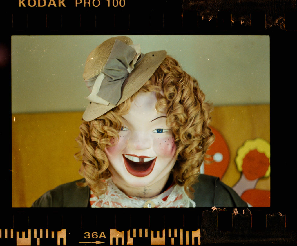 The photograph captures, with a precision that borders on reverence, the essence of a vintage clown doll. Its curly hair, meticulously crafted and eternally fixed in place, forms a halo around its porcelain face. Red lipstick adorns its lips in an exaggerated yet oddly endearing smile, one that seems to conceal as much as it reveals. The clown's attire—a beige hat adorned with a narrow ribbon and an elegant bow—adds a subtle grace to its otherwise garish appearance.

Encased within the familiar Kodachrome film border, the image radiates an innate sense of nostalgia. This frame is not merely decorative; it infuses the portrait with an unmistakable retro aesthetic that evokes memories of bygone eras and moments suspended in time. The clown doll stands alone but speaks volumes—a relic from another time, capturing both whimsy and melancholy in equal measure.
