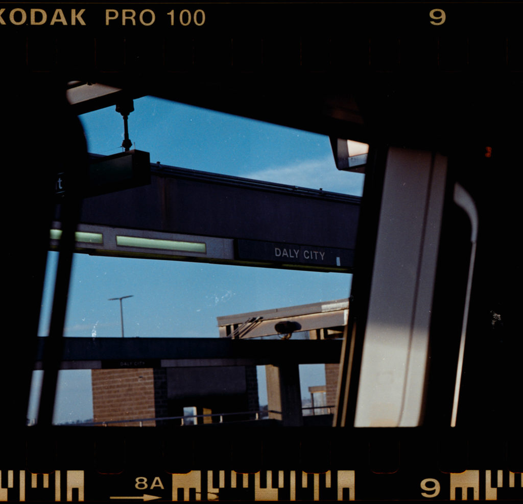 Through the smudged glass of the train window, a partial view emerges, fragmented by the reflections and the satin sheen of the interior. Beyond this scrim, a sign anchored to an outdoor platform comes into focus: "Daly City." The letters stand proud and purposeful under an unyielding glare of daylight. It refracts off every surface, charging even inanimate objects with a certain life.

The border around this moment is unmistakable—a Kodachrome embrace that imbues each shade and shadow with nostalgia. As steel wheels hum beneath me and strangers share silent company within these walls, I hold onto this fleeting glimpse framed in color—this intersection where journeys pause before they continue on their disparate paths.