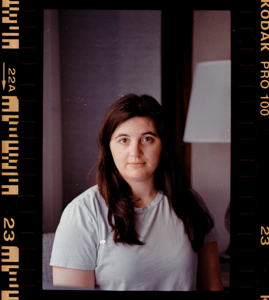 The photograph captures her essence with an uncanny precision, a woman with long, flowing dark hair cascading over her shoulders, wearing a light blue shirt that complements the deep brown of her eyes. She gazes directly at the camera, an unspoken narrative behind that steady look. The image is delicately bordered by film strips reminiscent of another era, hinting at the analog cameras that once held sway. In the softened blur of the background stands a lamp — its warm glow barely discernible, lending an intimate ambiance to this frozen moment in time.
