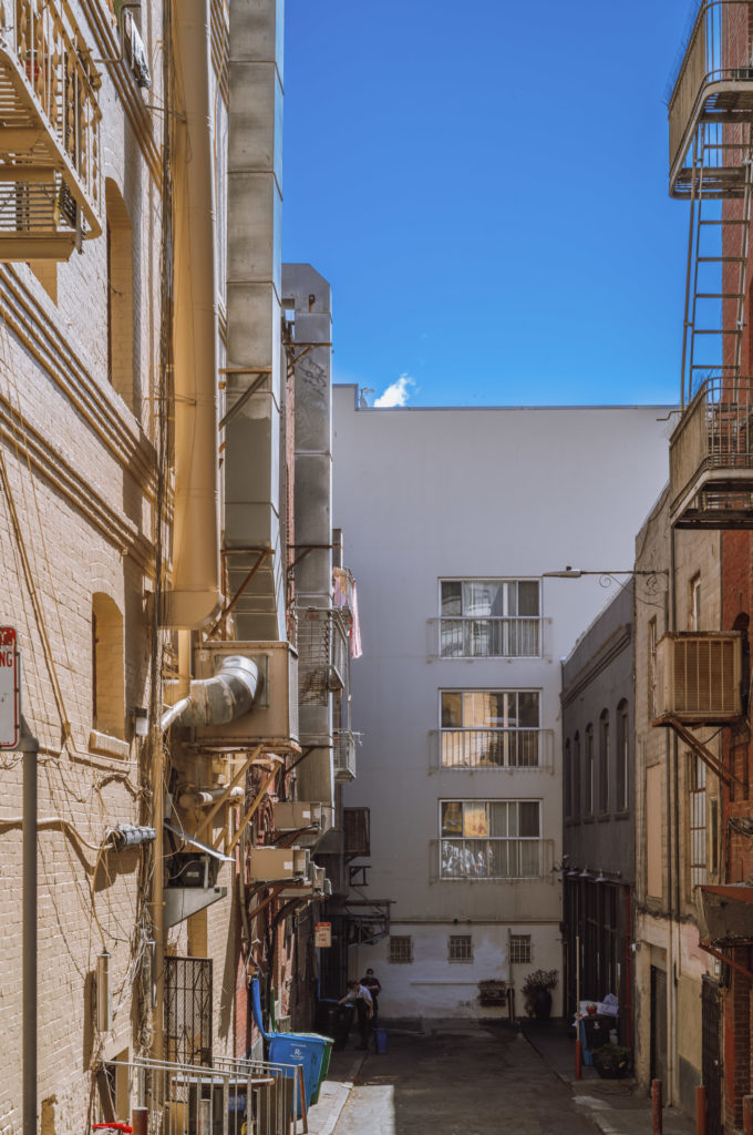 The narrow alleyway, flanked by timeworn brick buildings, whispered stories of an urban landscape where past and present coexisted in quiet harmony. Air conditioning units clung to the walls like reluctant guests, while a tangle of utility lines wove a chaotic but necessary web above. At the far end of this confined corridor stood a white building, its facade punctuated by several unblinking windows that seemed to gaze pensively at the clear, cerulean sky overhead.