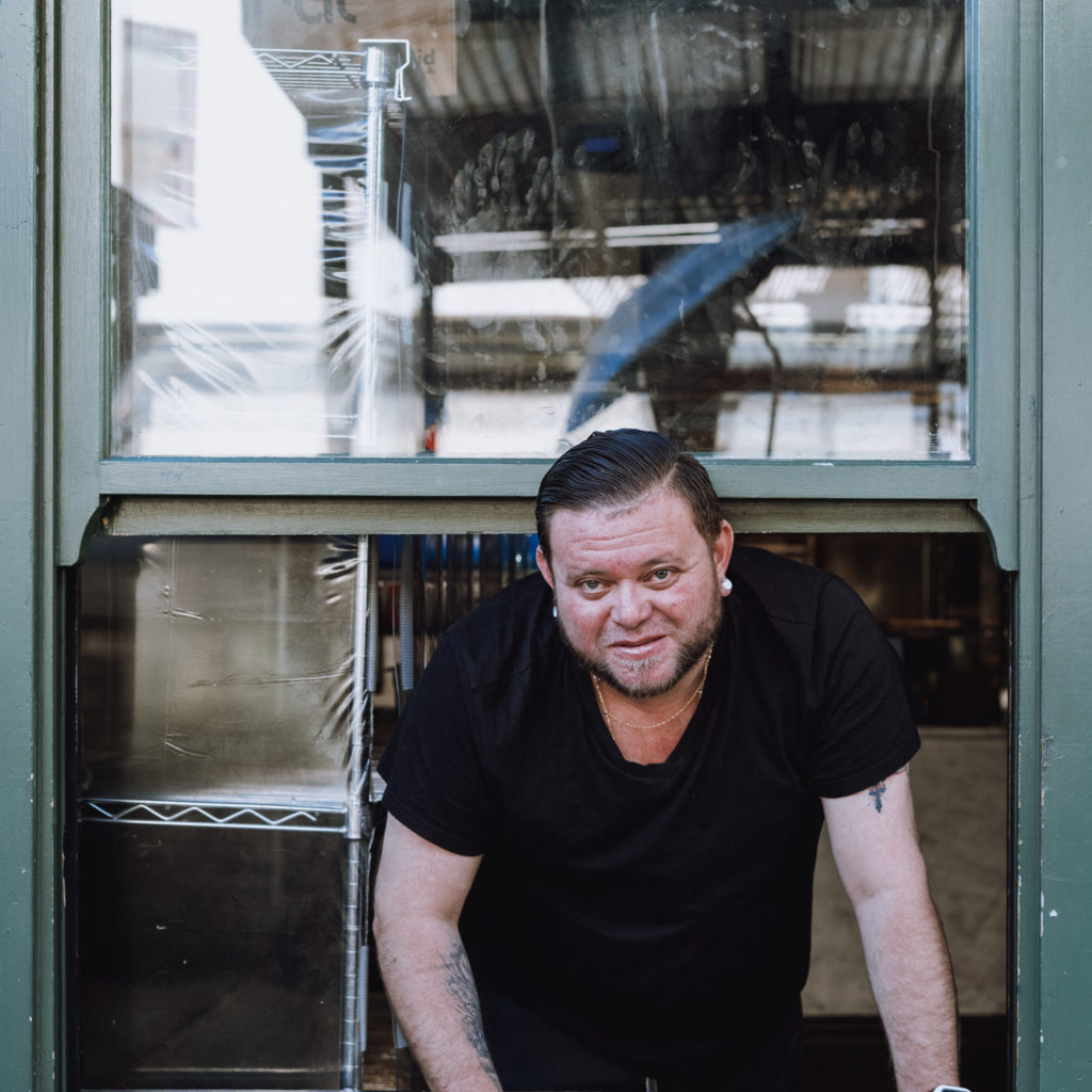 A man with short, dark hair and a neatly trimmed beard leaned out of the service window, his eyes scanning the room beyond. Clad in a simple black t-shirt that bore witness to countless shifts, his arms were adorned with intricate tattoos—stories inked into his skin. Behind him, the kitchen was a bustle of quiet activity: shelves lined with jars and spices, gleaming utensils hanging in anticipation, and pots simmering gently on well-worn stoves. Amidst this orchestrated chaos, he stood like an anchor—a steady presence in an ever-whirling world.