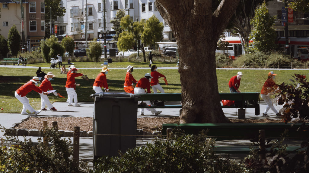 In the heart of the city park, a cluster of individuals, cloaked in garments of red and white, moved with a fluid grace. Their synchronized Tai Chi gestures unfurled like a silent dance beneath the watchful shade of an ancient tree. Shadows swayed as they embraced each deliberate motion. Beyond their tranquil enclave, the park buzzed with life: people strolled along winding paths, absorbed in conversations or solitude; others rested on benches, observing or lost in thought.

The backdrop was adorned with tall residential buildings which stood like sentinels against the sky's canvas. Trees framed this quiet urban sanctuary, their leaves whispering secrets to the wind. Each element—human and nature alike—wove together a tapestry that captured not just a moment but an essence of shared stillness amid life's perpetual motion.