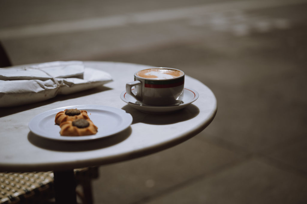 A small round table stood in quiet defiance on the bustling sidewalk, a solitary witness to the passage of time. Upon its surface rested a simple cup of coffee, steam curling lazily into the cool morning air, and beside it a delicate plate cradling a lone cookie. The cookie's edges were neatly crisped, an invitation to break the tranquility with each anticipated bite. Adjacent to this serene duo lay a wrapped parcel—its brown paper tied with humble twine—holding secrets or perhaps mundane necessities within.

The tableau was completed by its position at an outdoor café where life swept by in fleeting blurs of color and sound. The background thrummed with blurred figures streaming past, capturing snapshots of daily existence while remaining just beyond clear perception. Here, amidst life's perpetual motion, this little corner offered sanctuary—a brief pause where moments could be savored and thoughts given space to wander freely before rejoining the ceaseless currents flowing down the street.