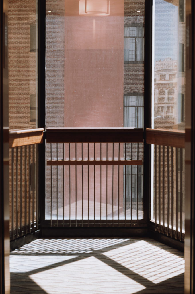 The balcony, a slender strip adorned with wooden railings and metal balusters, stood ensconced behind expansive glass panes. The sunlight seeped through, scattering intricate shadows across the floor like an artist's careful brush strokes. Beyond the transparent barrier loomed a brick building punctuated by numerous windows, each framing its own quiet tableau of urban life.